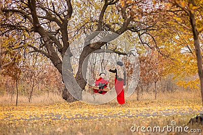 Beautiful mother in a long dress is swinging on a hinged swing little daughter in a red coat in an autumn garden near an old gnarl Stock Photo