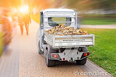 Small electric truck removing fallen leaves in body at autumn city park. Municipal urban services using ecology green vehicle Stock Photo