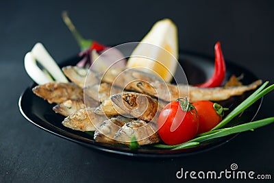 Small fried herrings with vegetables on a black plate on a black background. Stock Photo