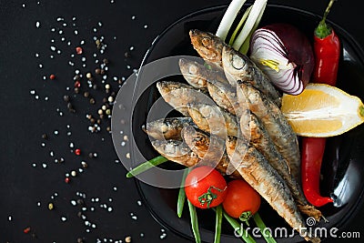 Small fried herrings with vegetables on a black plate on a black background. Fish appetizer. Stock Photo