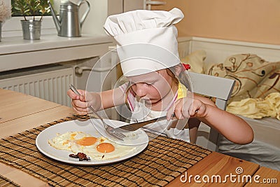 Small girl has a snack. Little girl eats ham and eggs. Cute girl wears chef costume. Family and childhood concept. Stock Photo