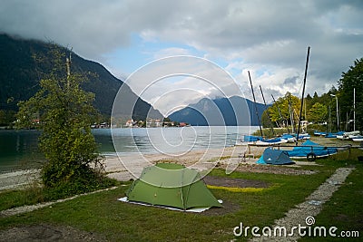 Small green tent and boats next to beach on campground camping Walchensee in the alpine upland with lake and mountain Herzogstand Stock Photo