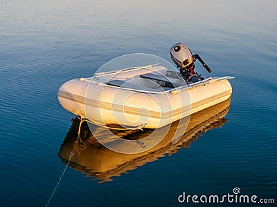 Small inflatable dinghy used as yacht tender with outboard motor moored alongside a yacht on a calm, glassy sea at sunset Stock Photo