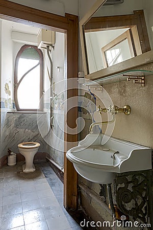 In a small room there is the toilet with the broken toilet seat, outside the door is the sink with the faucet in gold Stock Photo