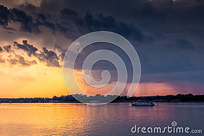 A small ship is sailing along the river against the backdrop of the sunset. Rain clouds in the sky Stock Photo