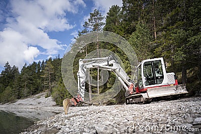 Small shovel digger standing on gravel next to lake and forest Stock Photo