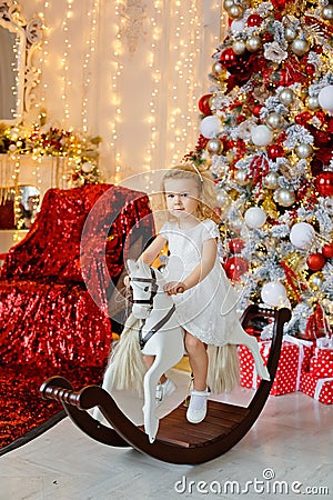 A small smiling curly-haired blonde girl in a white dress is sitting on a rocking horse under a Christmas tree against the Stock Photo