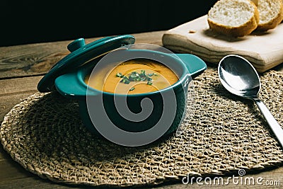 Small turquoise pot with mashed vegetables, olive oil and chives on a wicker mat with a spoon and a wooden board with slices of Stock Photo