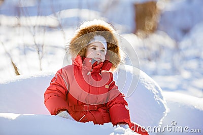 Small very cute girl in a red suit with fur hood sits in the snow in winter forest background Stock Photo