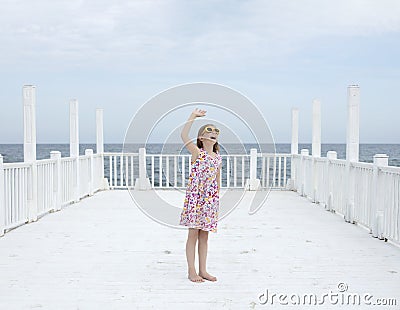Small 8 years old pretty cheerful girl in a dress with a floral print and in yellow sunglasses stands on a wooden white pier Stock Photo