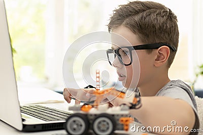 A smart boy builds a robot car using a laptop near the window at home. The child is learning coding and programming Stock Photo