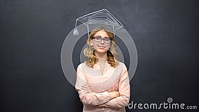 Smart girl stands against academic cap picture on blackboard, education program Stock Photo