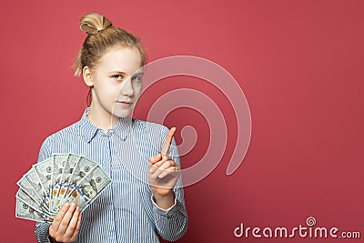 Smart happy cunning young girl showing money us dollars and pointing finger up on pink background Stock Photo