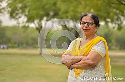 Smart senior north Indian woman standing, posing for the camera with hands crossed in a park wearing yellow salwar kameez in Stock Photo