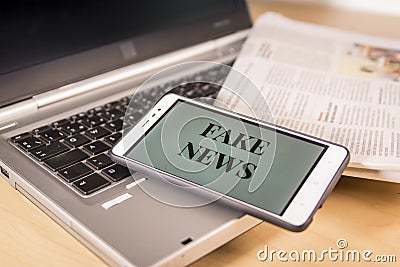 Smartphone with Fake News words on screen over a newspaper and a laptop. Fake news, HOAX concept Stock Photo