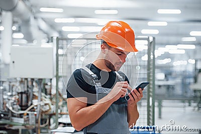 Smartphone in hands. Industrial worker indoors in factory. Young technician with orange hard hat Stock Photo