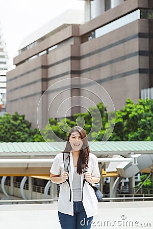 Smiled young Asian woman in white shirt carrying a backpack standing on office building square with office building background Stock Photo