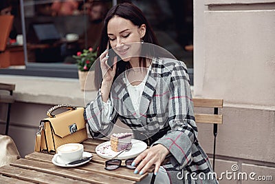 Smiled young pretty woman having phone talk while eating cheesecake with cappuccino in cafe. Stock Photo