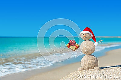 Smiley sandy snowman at beach in christmas hat with golden gift. Stock Photo