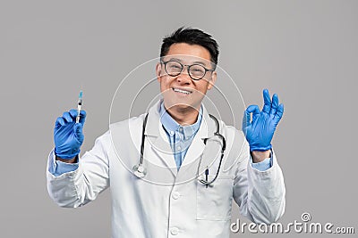 Smiling adult asian doctor in white coat, glasses, protective gloves hold syringe, show ok Stock Photo