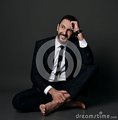 Smiling adult bearded man in black office suit sits barefooted on floor with legs crossed holding hand at head Stock Photo