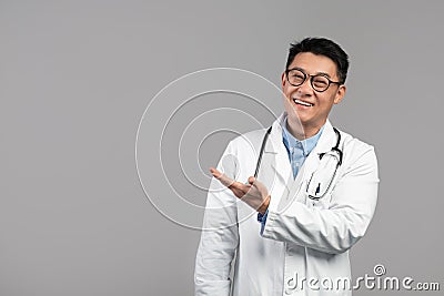Smiling adult korean man therapist in white coat, glasses with stethoscope shows hand on empty space Stock Photo