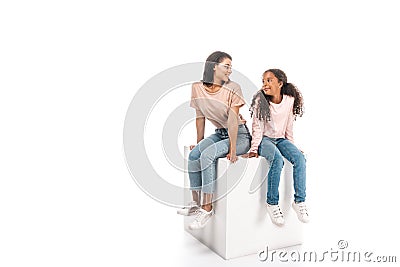 African american mother and daughter looking at each other while sitting on white cube on white background Stock Photo