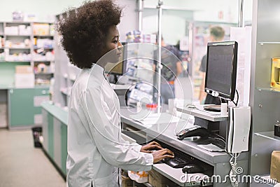 Smiling African American woman clinical pharmacist typing on computer keyboard, while standing behind the counter of Stock Photo