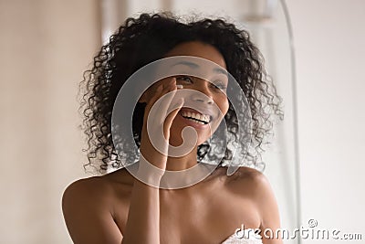 Smiling biracial woman apply under eye cream looking in mirror Stock Photo