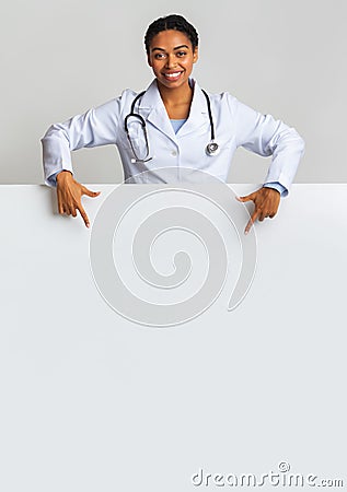 Smiling afro female intern doctor pointing at blank advertisement board Stock Photo