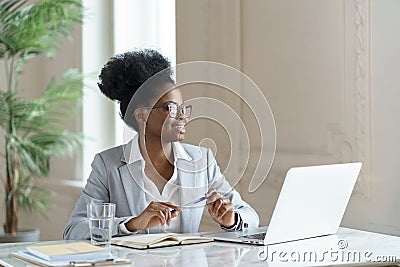 Smiling Afro woman in blazer wear glasses working at laptop computer at home office, looking away. Stock Photo