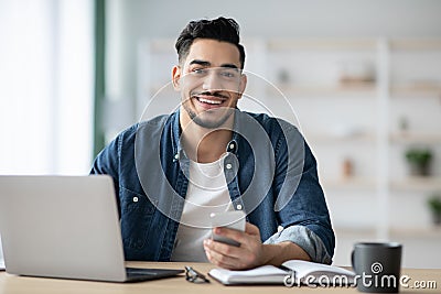 Smiling arab guy using smartphone while sitting at workdesk Stock Photo