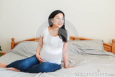 Smiling Asian Chinese pregnant woman sitting on bed touching belly. Expecting young lady in white plain t-short and blue jeans at Stock Photo