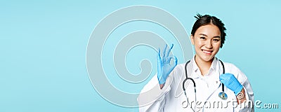 Smiling asian doctor, female physician touching stethoscope, showing okay, ok sign in approval, blue background Stock Photo
