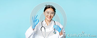 Smiling asian doctor, female physician touching stethoscope, showing okay, ok sign in approval, blue background Stock Photo