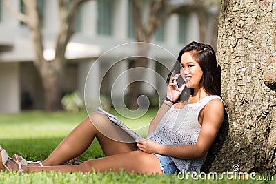 Smiling Asian girl talks on cell phone while studying under a tr