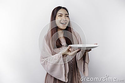 A smiling Asian Muslim woman is fasting and hungry and holding and pointing to a plate Stock Photo
