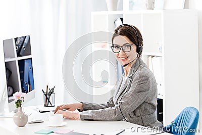 Smiling attractive call center operator in glasses and headset using computer at table in office, looking Stock Photo