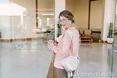 Smiling attractive girl entering big glass door into office, hotel, business centre. Wearing stylish glasses, gray pants Stock Photo