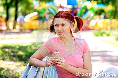 Smiling young woman with pigtails hairstyle and pink shirt sitting on bench in park and holding ice-cream in waffle cone Stock Photo