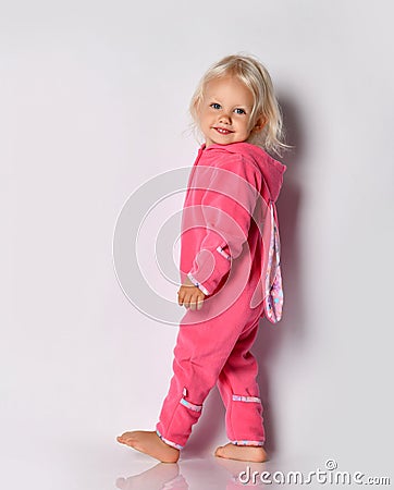 Smiling barefoot cute blonde baby girl in pink warm comfortable jumpsuit with hood and bunny ears walks passes by camera Stock Photo