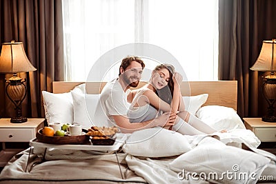Smiling bearded man being happy to spend free time with his female lover, sit together in comfortable bed in hotel room Stock Photo