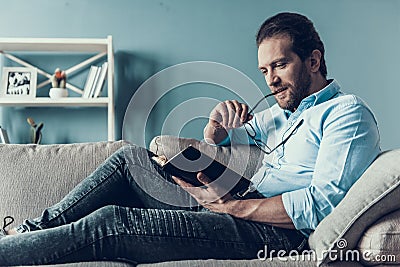 Smiling Bearded Man Reading Book on Gray Sofa. Stock Photo