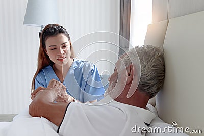 Smiling beautiful caring female doctor holding hand of male senior patient who lying in hospital bed. Nurse takes care elderly man Stock Photo