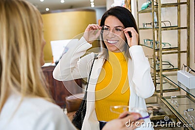 Smiling beautiful woman choosing and showing eyeglasses to attractive young oculist in optic shop Stock Photo