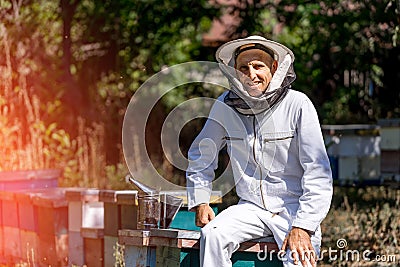 Smiling beekeeper in white uniform. Man sitting near work equipment and beehives. Stock Photo