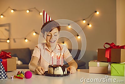 Smiling birthday boy in festive cap sitting and looking at birthday cake with candles Stock Photo