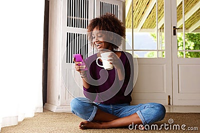 Smiling black woman sitting on floor at home with cell phone Stock Photo