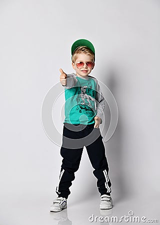 Kid boy in grey sweatshirt, black sport pants with stripes and white sneakers stands gesturing thumb-up Stock Photo