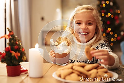 Smiling blonde girl holding mug with cacao and cookie, looking a Stock Photo